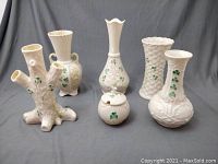 Full lot of six assorted Belleek vessels displayed on a gray backdrop showing varied shapes and green shamrock decoration.