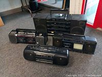 Full view of multiple vintage portable radios and cassette player boomboxes arranged on carpeted floor, showing condition and general appearance of items for repair.