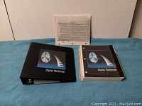 Photo showing the black 3-ring binder labeled Digital Performer, sealed software packet, and loose-leaf information sheets on a blue fabric surface.