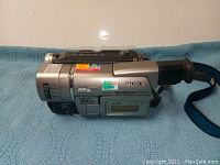 Side angled photo of silver Sony Handycam Vision video camera showing the brand, model labels, hand strap, and LCD display.