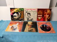 Photo shows 6 vinyl record LP album covers arranged in two rows. Front row includes 30 Hits of the 50's, Judy Holliday, Sue Wilts, Love Story soundtrack, My Fair Lady, Calypso Heat Wave. Covers show light wear and vintage artwork.