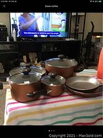 Set of five copper-colored Farberware nonstick cookware pieces, three pots with lids and two frying pans, displayed on a striped cloth in front of a TV.