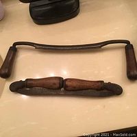 Upper image shows two vintage sling blades laid out on a smooth surface with wooden handles and curved metal blades, one fully extended and one partially folded.