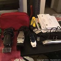 Four hair clippers arranged on a black surface alongside a red carrying case and some papers, cords wrapped around clippers visible