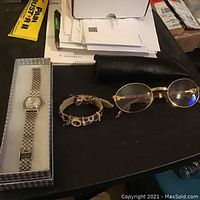Lot showing Nixon ladies watch in box, charm bracelet, and round gold eyeglasses on black surface with papers in background