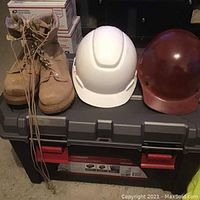 Full view of tan work boots, white hard hat, and brown hard hat placed on a plastic storage container.