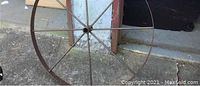 Close-up photo of large metal spoked wheel from vintage spinning wheel showing rust and weathering; wheel has wooden handle attached.