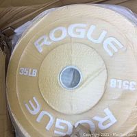 Top view of yellow Rogue bumper weight plate in original packaging, displaying brand and weight markings.
