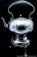 Front view showing rounded kettle body, engraved floral patterns, curved spout, and arched handle above the kettle attached by scroll supports. Warmer base ornate and detailed.