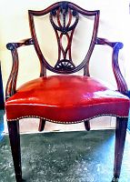 Front view of antique armchair showing carved wooden backrest, curved armrests and red leather seat with nailhead trim.