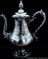 Photo showing antique silver-plated coffee pot with detailed embellishments on handle, spout, and lid finial, mounted on pedestal base.