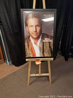 Framed signed Kevin Costner Bull Durham poster displayed on wooden easel against a black curtain.