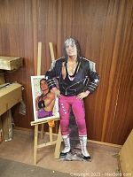Full view of Bret Hart cardboard cutout standing with arms on hips in front of wood panel wall, with additional print on easel nearby