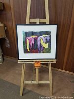 Framed colorful abstract painting on an easel against wood paneled wall, with colorful figures in purple, yellow, and green.