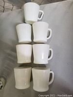 Seven white vintage milk glass coffee mugs arranged in two rows, varying slightly in shape and size, on a gray surface.