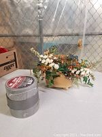 Photo showing artificial floral arrangement in ceramic pot with 3 rolls of grey duct tape stacked on the left side.
