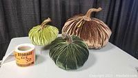 Set of three velvet pumpkins in green and brown shades, deflated and placed on a table, showing size comparison alongside a roll of tape.