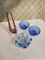 Overview photo of lot showing tall pink and clear glass vase, two blue glass custard cups, and blue-yellow candy bowl on white table.