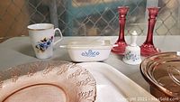 Photo shows assortment of blue cornflower Corning Ware dishes, pink glassware such as candlesticks and plates, and other decorative and functional kitchen items.