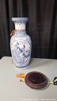 Large ceramic vase with intricate peacock and floral design and wood stand base on a white surface.