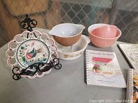 Overview of lot items including hand painted Portugal bird plate, metal stand, Fire-King wheat dish, pink Pyrex bowls, vintage Canadian jar, and cookbooks.