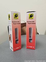 Front and side view of two boxed J-Jati electronic mill pepper grinders on table showing product image and brand logo.