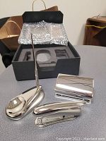 Desk set items arranged outside of the box on a grey surface, showing pen with stand, puncher, stapler, and paper clip holder.