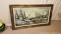 Front view of large framed mountain landscape painting showing snow-covered trees, mountains, stream and waterfall in a decorative gold frame.