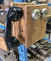 Side view of the antique wall phone showing oak casing, black handset, and metal ringer bells on front.