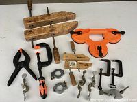 Assortment of clamps arranged on white surface showing wooden bar clamps, plastic suction clamp, plastic and metal clamps of various sizes and types.
