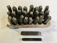 Top view of a wooden holder containing metal stamps with numbers and letters, arranged side by side, showing wear and some rust.