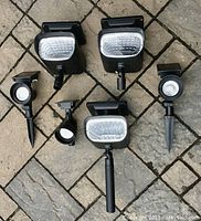 Six solar spotlights laid on stone pavement: three large rectangular floodlights and three smaller circular spotlights, all with black housings and solar panels on top.
