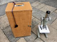 Vintage Tasco Student 100X microscope next to its vintage dovetailed wooden carrying case, outdoors on concrete ground.