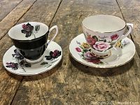 Two teacup and saucer sets on wooden surface, black and red rose Royal Albert and colorful floral Royal Sutherland.