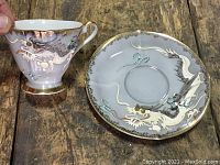 Teacup held next to saucer showing detailed white dragon design with moriage on bluish grey background with gold trim