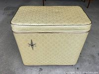 Closed view of rectangular vintage MCM clothes hamper showing yellow quilted vinyl exterior with diamond and cross pattern and metallic star emblem on front