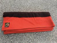 Folded red blanket with black stripe and Trapper Point label showing next to label patch image of Native American in traditional headdress.