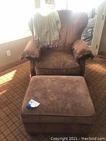 Front view of Marshf II upholstered wingback chair with a brown textured fabric, brass nailhead trim, and a matching ottoman placed in front. Blanket draped over chair is not included.