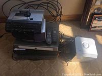 Photo showing stacked electronic devices including Sony and GoVideo VHS/DVD combo players, TiVo box, Brookstone clock radio, Sanyo recorder, and Dell subwoofer on carpeted floor.