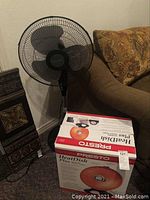 Boxed Presto HeatDish Plus parabolic heater shown on the floor near the Holmes fan and sofa, box is intact and shows photo of the orange parabolic heat dish.