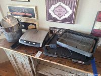 Photo showing George Foreman grilling machine, electric griddle, roasting pans, and pots arranged on wooden cabinet under wall decorations