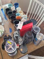 Overview of lot on table showing red Nespresso machine, NutriBullet blender, various ceramic mugs, coffee pots, and tumblers