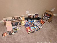 Photo showing an assortment of DVDs, CDs, and VHS tapes spread on floor with some VHS tapes in cardboard boxes.