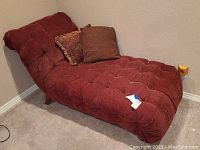 Side angled view of red chaise lounge showing tufted upholstery and two pillows against beige wall on carpeted floor.