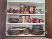 Three shelves of cabinet holding various colorful ceramic dishes including mugs, pitchers, bowls, and plates in red, brown, and beige hues.