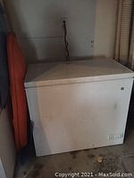 White GE chest freezer placed against wall with closed top lid and power cord plugged in, floor beneath shows minor dirt and object next to freezer.