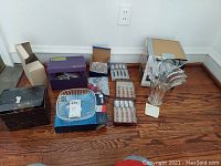 Wide shot of lot items arranged on floor including boxed and unboxed bowls, wine glasses, stationery, and a large crystal vase.