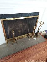 Overall view of fireplace screen, brass andirons, and tool holder with tools arranged near fireplace.