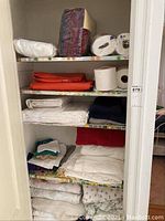 Closet shelf with various linens including towels, sheets, folded white and colored fabrics, some paper towels.