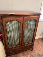 Front view of wooden cabinet showing two doors with decorative wire mesh and fabric curtain behind, and lower drawer with handles.